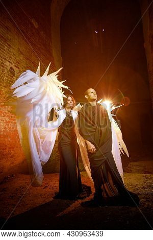 Couple In Dark Clothes In An Abandoned Castle With Old Brick Walls. Man In Black Dress And Woman Wit