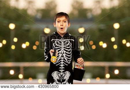 halloween, holiday and trick-or-treating concept - excited boy in black costume of skeleton with candies and flashlight over garland lights at roof top party background
