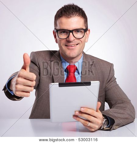 young business man sitting at the desk with a tablet in his hand and showing the thumb up gesture while smiling for the camera. on a gray background
