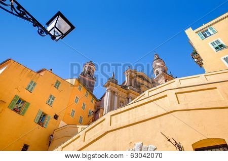 Yellow and orange residential houses and Saint Michel Archange basilica under clear blue sky in Menton, France.