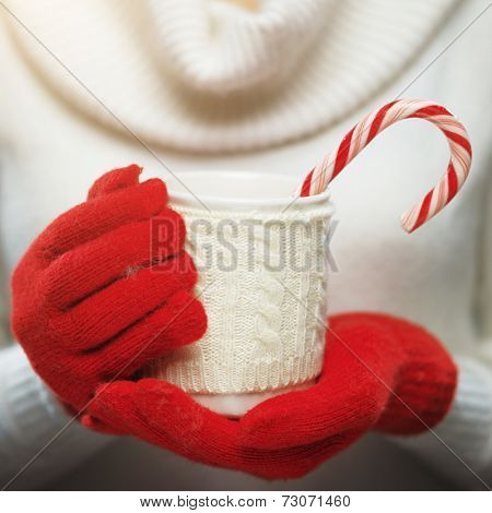 Woman holds a winter cup close up. Woman hands in woolen red gloves holding a cozy mug with hot cocoa, tea or coffee and a candy cane. Winter and Christmas time concept.  