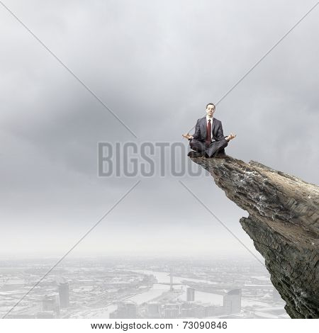 Young businessman sitting lotus pose on edge of rock mountain