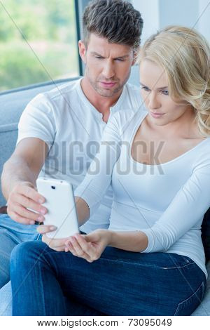 Couple reading a text message on their mobile phone with consternation frowning at the screen as they sit together on a sofa