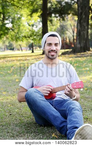 Portrait of young handsome man playing the ukelele in a park. Outdoors.