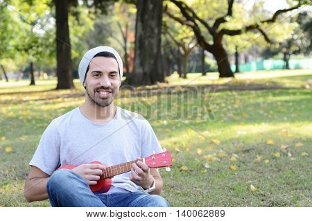 Portrait of young handsome man playing the ukelele in a park. Outdoors.