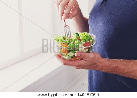 Man eat healthy lunch in modern interior. Unrecognizable profile male torso in blue t-shirt, hand with fork, near window with vegetable salad in bowl, diet food concept. High key image