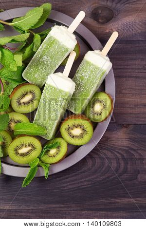 Homemade frosty kiwi popsicles on a plate with mint and kiwi slices on a dark wooden background. Top view with copyspace