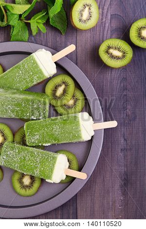 Homemade kiwi popsicles on a plate with mint and kiwi slices on a dark wooden table. Top view with copyspace