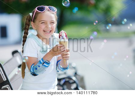 A cute little girl with two long braids, wearing sun glasses in a pink frame with glass in the shape of hearts, wearing gold earrings, wearing a white blouse and blue shorts holding an ice cream in waffle cone sitting on a bench in the park after rollerbl