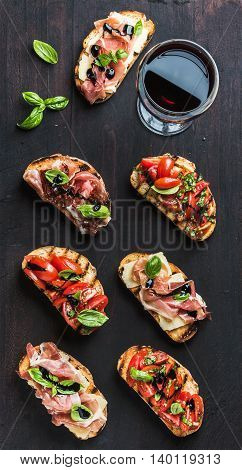 Brushetta set with glass of red wine. Variety of small sandwiches with prosciutto, tomatoes, cheese, herbs and balsamic creme on dark wooden background, top view