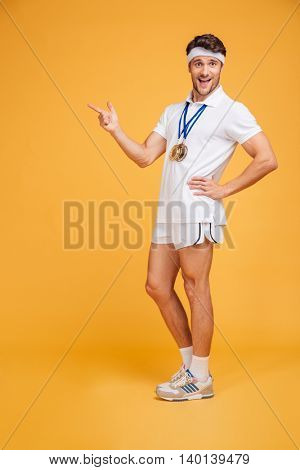 Smiling young spotrsman with three medals sranding and pointing away over yellow background