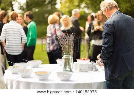 Banquet lunch break at conference meeting on hotel terrace. Assortment of food and beverage. Unrecognizable people discussing over food in background.