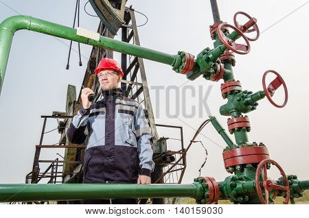 Oilfield worker near wellhead wearing red helmet and work clothes holding the radio. Pump jack background. Oil and gas concept.