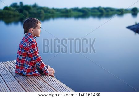 boy at dusk sits on the docks and looks afar. pensive boy in a plaid shirt sitting on a wooden pier near the water. side view. empty space for your text