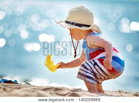 one year old boy playing at the beach in straw hat. Child with a paper ship at sea