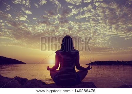 Silhouette of a young woman practicing yoga on a rocky shore in Syvota bay Greece. Cloudy sky Ionian sea and a beautiful sunset in the background.