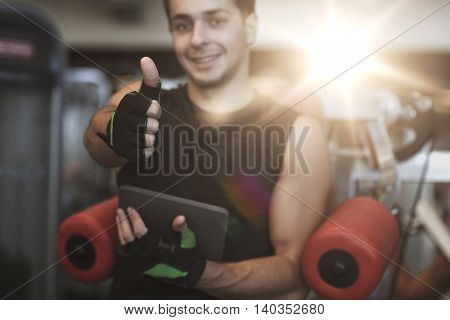 sport, bodybuilding, technology, gesture and people concept - happy young man with tablet pc computer showing thumbs up in gym