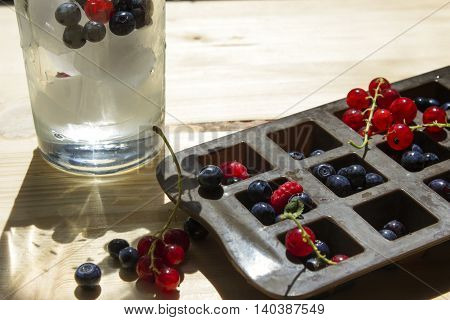 Wild berries in an ice cube tray and glass with cold drink on a wooden table.