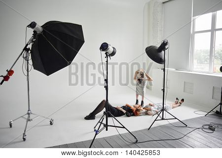 Girl photographer photographing fashion models in a clear coat with a pomegranate mask on the face and black clothes lying on white floor in Studio