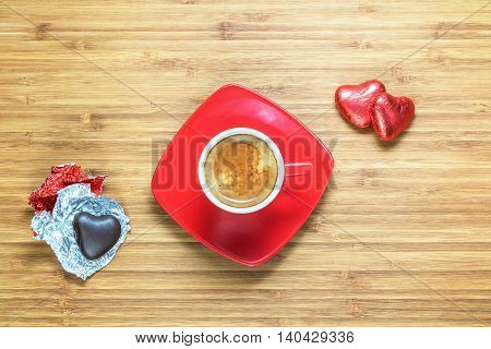 Heart shaped sweets wrapped in a bright red foil lying on a wooden texture with red cup of coffe near it. Background for romantic themes.