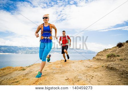 Couple runners running with backpacks on rocky trail at seaside and mountains. Young woman and man trail running on mountain path looking at beautiful inspirational landscape view.