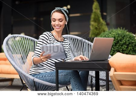 Pleasant work. Cheerful charming content woman sitting at the table and listening to music while using laptop