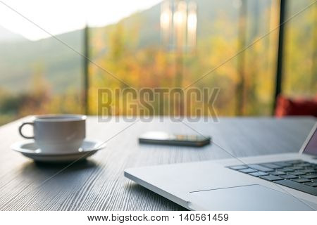 Computer Coffee Mug and Telephone on black wood table green flora beyond large windows on background. Focus on front part of laptop and touchpad