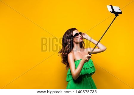 Portrait of young model in green dress and sunglasses posing while making selfie against yellow background.