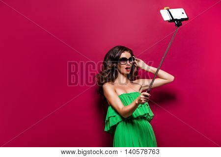 Portrait of young model in green dress and sunglasses posing while making selfie against red background.