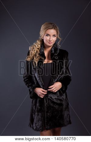 Fashionable young woman with long blonde hair smiling at camera while wearing black fur coat. Studio shot.Isolated.