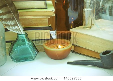 Retro still life with vintage things: old books pen with ink jar glass bottles burning candle and a smoking pipe. Photo with toning.
