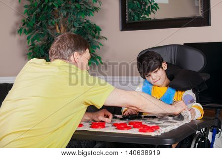 Young biracial disabled boy in wheelchair playing checkers with father at home