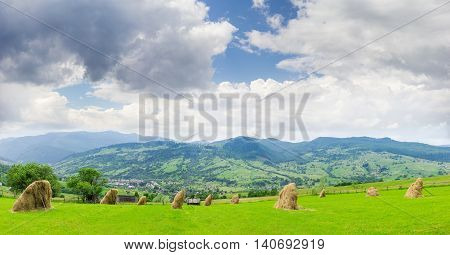 Mountain landscape with hayfield and hay which is dried on the foreground mountain village in the valley on the background of mountain ranges and sky with clouds