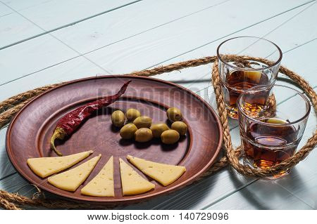 Two whiskey glasses and a plate of olives cheese and pepper on a blue wooden background.