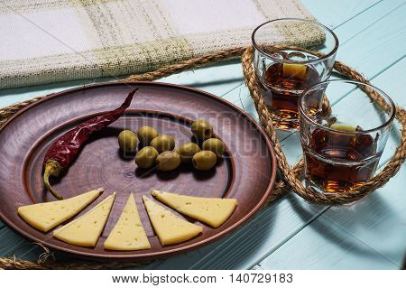 Two whiskey glasses and a plate of olives cheese and pepper on a blue wooden background.