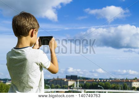 Boy with mobile phone. Child taking photo with his smartphone. Beautiful sky and city background. Back view. Technology leisure gaming concept