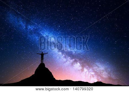 Night landscape with Milky Way. Silhouette of a standing man with raised up arms on the top of mountain. High rocks mountain peak. Beautiful Galaxy. Universe. Blue night starry sky and city lights