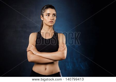 Young beautiful sportive girl posing with crossed arms over dark background. Copy space.