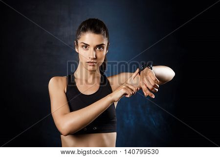 Young beautiful brunette sportive girl pointing at watch over dark background. Copy space.