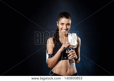 Young beautiful brunette sportive girl holding water over dark background. Copy space.