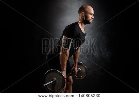 Young powerful sportsman in heaphones training with barbell over black background. Copy space.