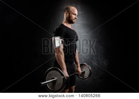 Young powerful sportsman in heaphones training with barbell over black background. Copy space.