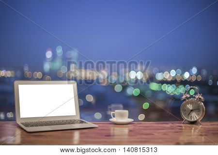 Front view of creative designer desktop with blank white laptop silver alarm clock and coffee cup on blurry night city background. Mock up