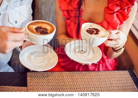 Engagement Ring Under A Cup Of Coffee. Man And Woman Holding It, With Hearts In Cafe