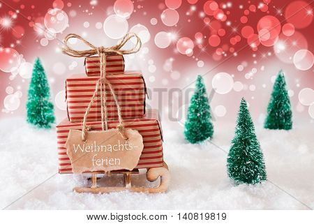 Sleigh Or Sled With Christmas Gifts Or Presents. Snowy Scenery With Snow And Trees. Red Sparkling Background With Bokeh Effect. Label With German Text Weihnachtsferien Means Christmas Break