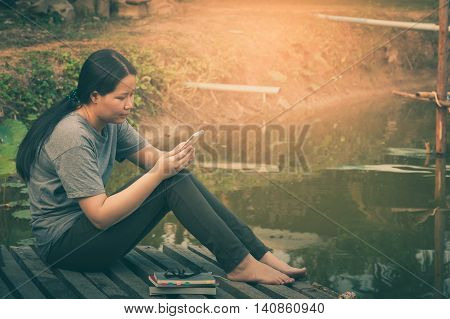 Weekend morning lifestyle. Young woman using her mobile phone seriously while sitting outdoor beside lake in morning time. Freelance working and phone addiction concept with vintage filter effect