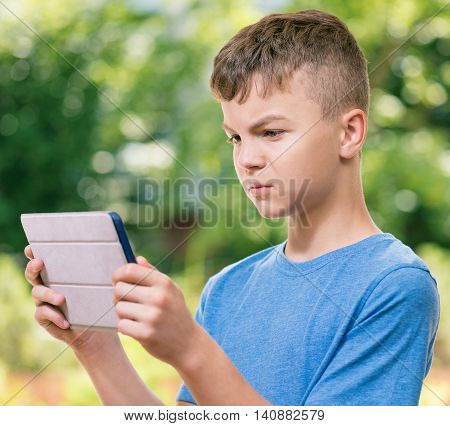 Outdoor portrait of serious teen boy 12-14 year old with tablet pc watching video in summer park