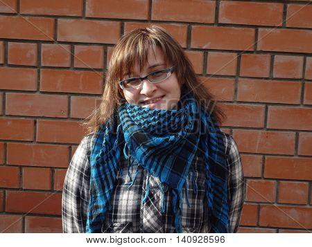 Happy girl in glasses and plaid jacket and scarf stay about brick wall