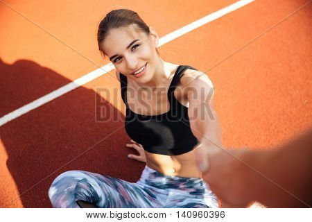 Smiling attractive young woman athlete getting up and taking somebody hand on stadium