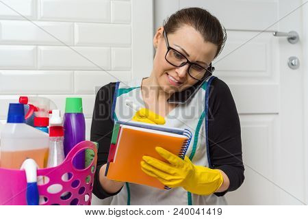 Housemaid Woman Is Cleaning. Woman In Yellow Rubber Gloves, Talking On Phone Writing In Notebook